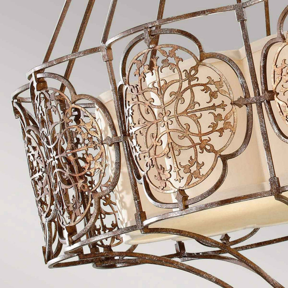 Close-up of the Marcella 4 Light Chandelier - Bronze, showcasing an ornate, vintage metal lampshade with intricate, symmetrical floral patterns encircling a cream-colored fabric inner shade. The metal boasts a rustic bronze finish with an elegant touch, enhancing its luxury lighting appeal.