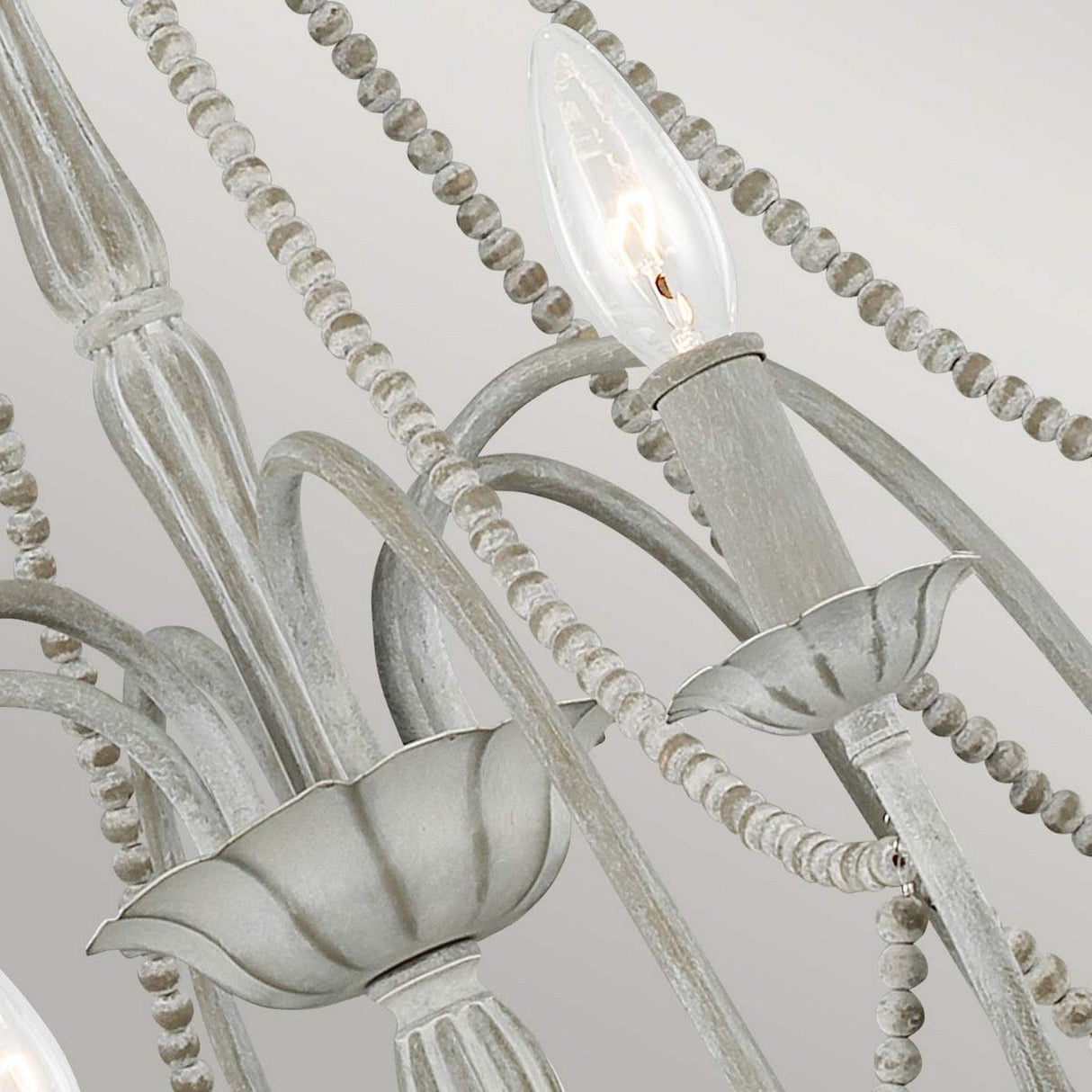 A detailed view of the Maryville 6 Light Chandelier in Washed Grey highlights its rustic elegance, with candle-like light bulbs and decorative beads. The metal frame boasts a weathered finish, embodying French Country style, and the design includes floral-shaped elements and gracefully curved arms.