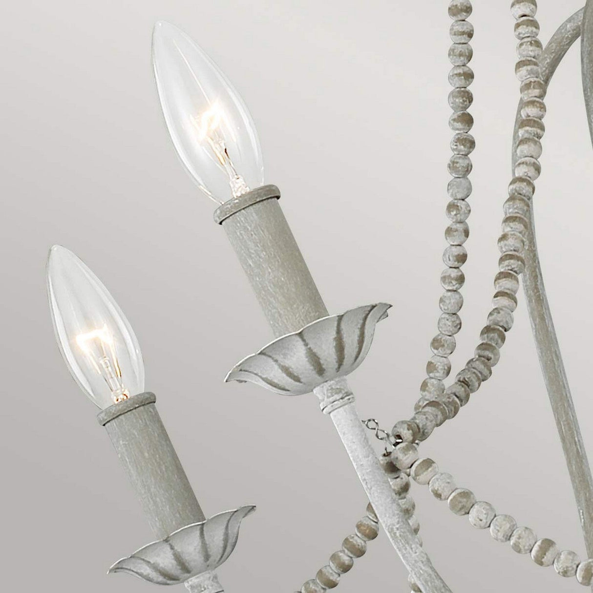 A close-up of the Maryville 6 Light Chandelier, showcasing its rustic elegance with two lit candle-style bulbs. This fixture embodies French Country style with a distressed white finish and decorative beaded chains, set against a soft gray background.