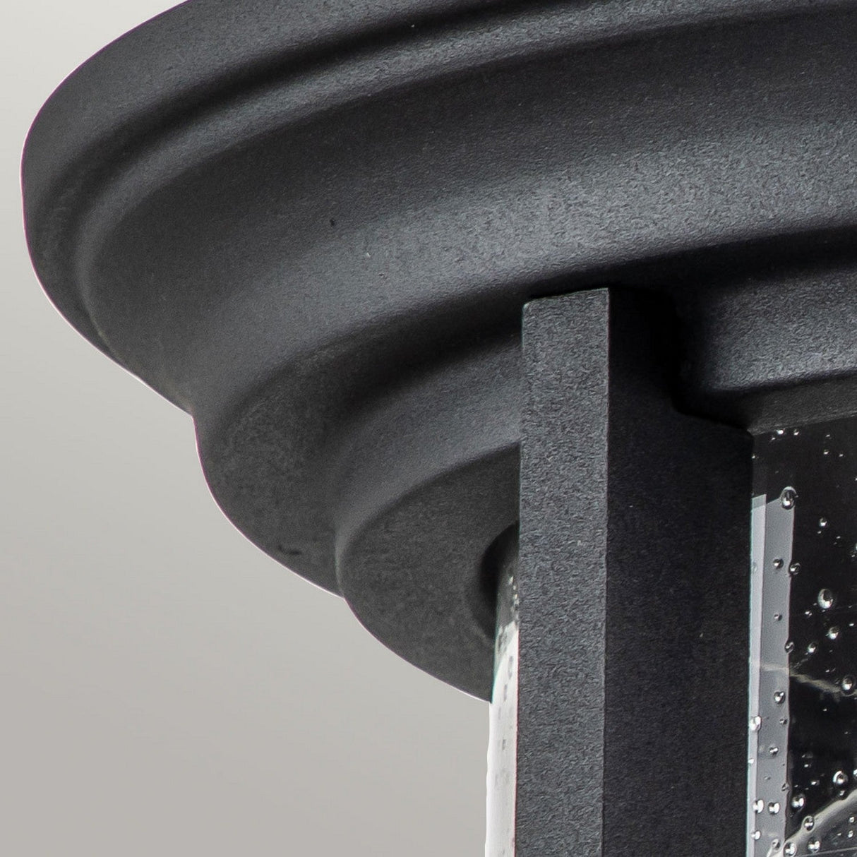 Close-up of the Merrill 2 Light Outdoor Flush in black, featuring a dark, textured lantern design with a curved metal top and clear seeded glass. Its modern and industrial aesthetic highlights the detailed craftsmanship against a light gray backdrop.