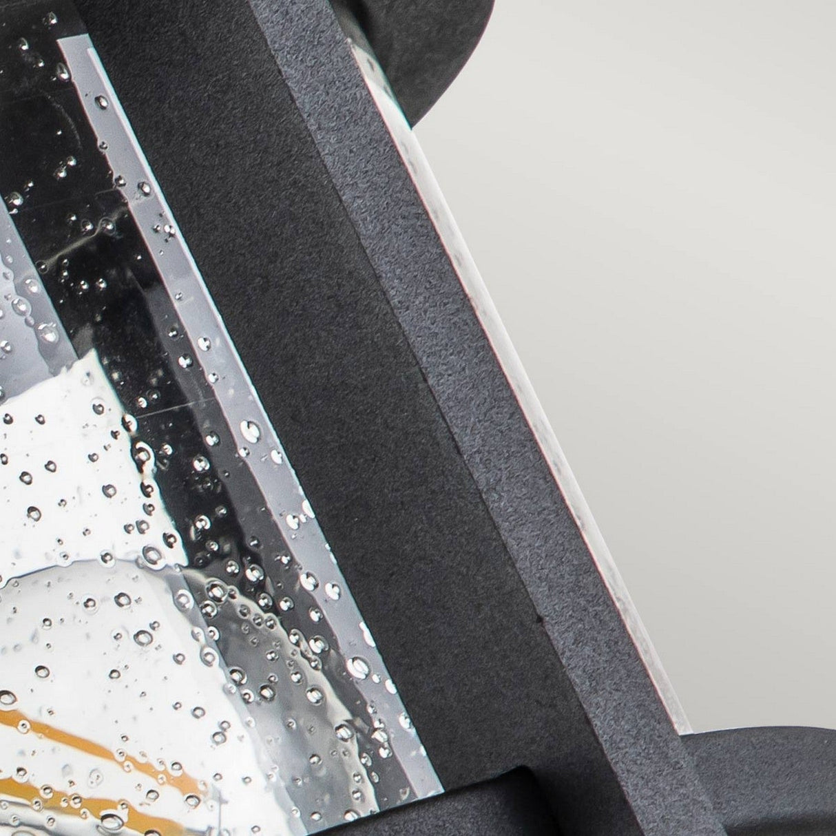 A close-up showcases a rain-soaked glass panel adorned with water droplets, encased in the frame of the Merrill 2 Light Outdoor Flush - Black. The clear seeded glass artfully softens the light gray backdrop, accentuating its charm against the textured black structure.