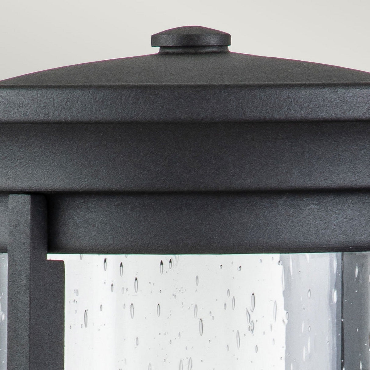 Close-up of a dark metal outdoor wall lantern, similar to the Merrill 1 Light Small Outdoor Wall Lantern - Black, showcasing a rain-speckled seeded glass panel. The lantern's top has a rounded design with a small, flat cap. The droplets' texture is visible on the glass against the soft gray background.