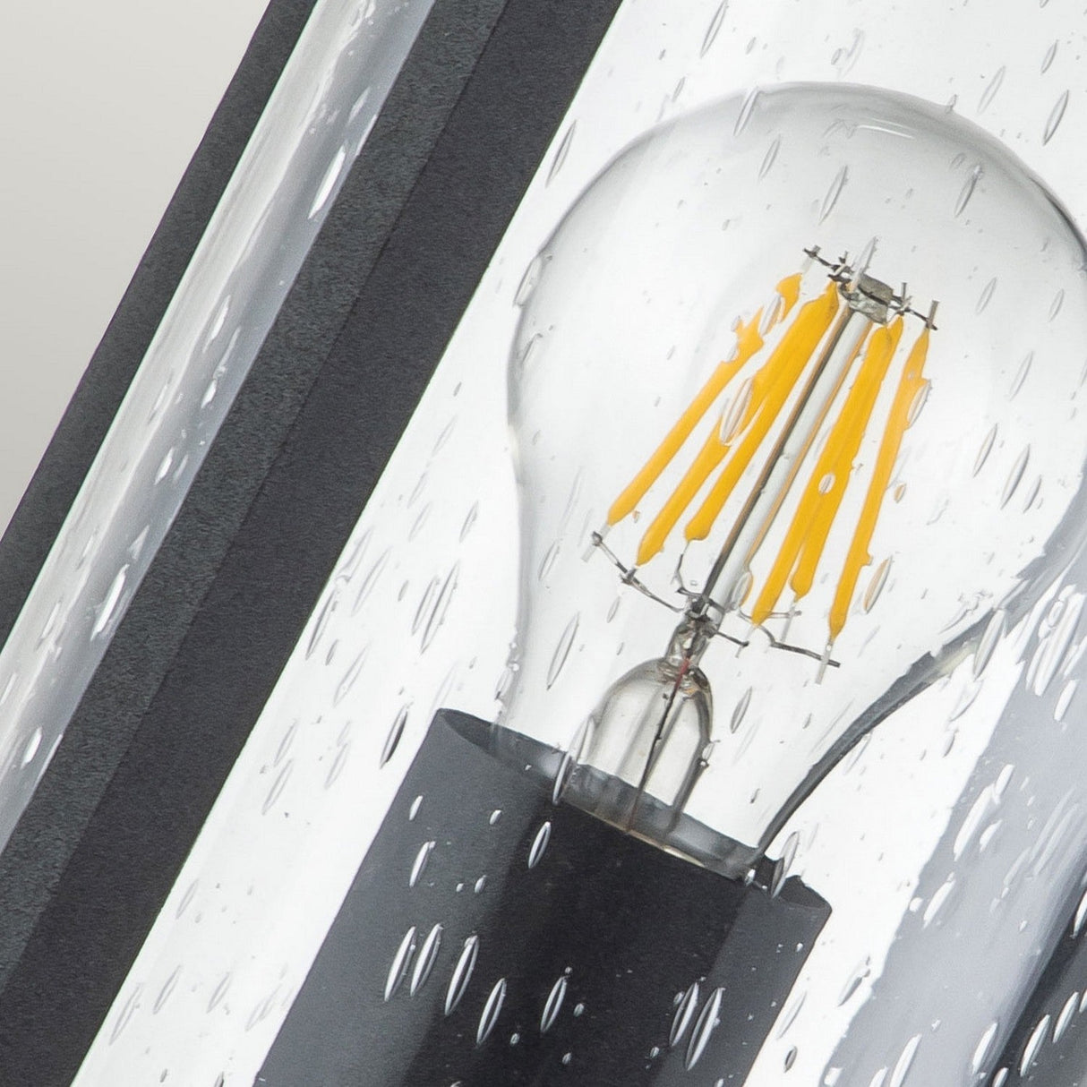 A close-up of the Merrill 1 Light Small Outdoor Wall Lantern - Black, reveals a light bulb inside seeded glass with water droplets on the surface. The visible filaments and black base enhance its resemblance to an outdoor wall lantern in a moist setting.