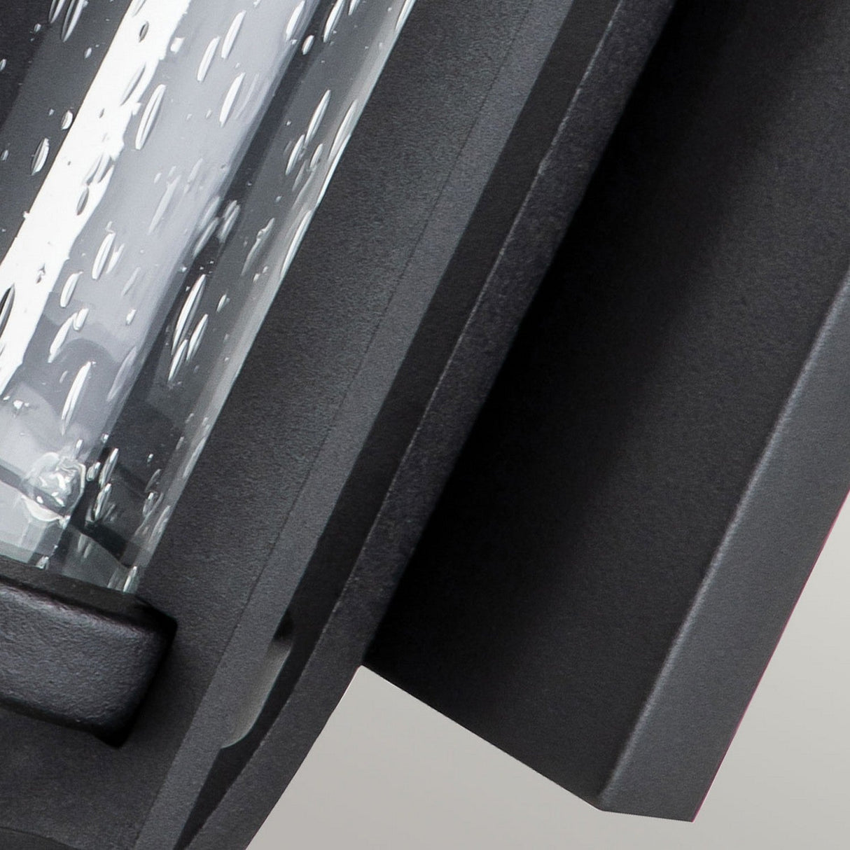 Close-up of a contemporary outdoor wall lantern featuring seeded glass with water droplet patterns. The Merrill 1 Light Small Outdoor Wall Lantern - Black highlights a sleek frame that showcases modern design elements.