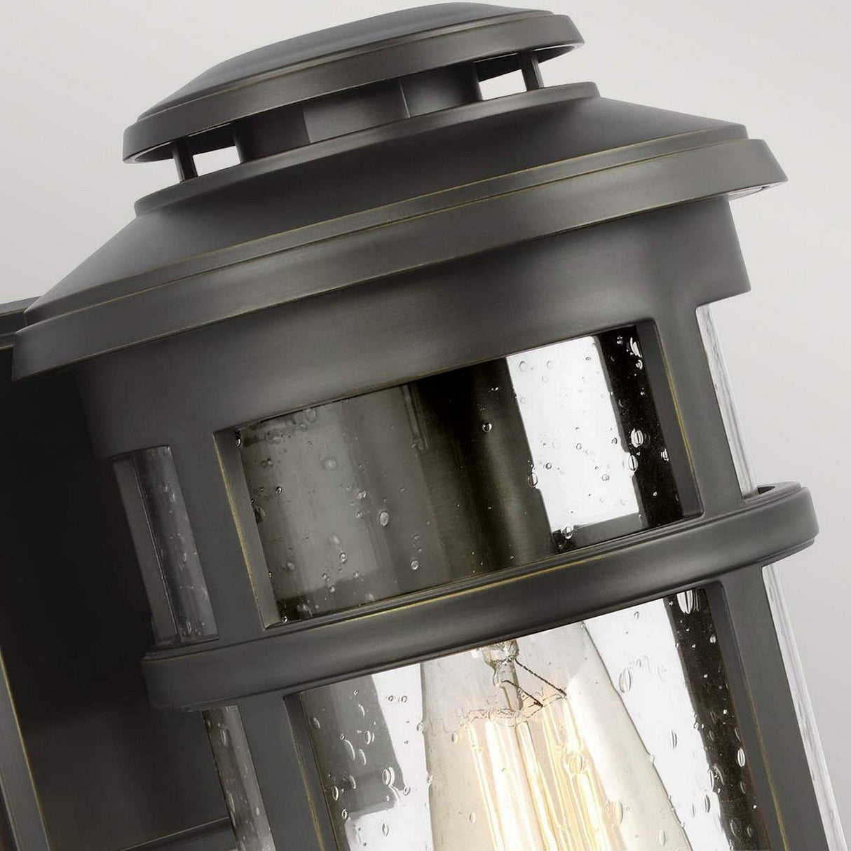 Close-up of the Newport 1 Light Outdoor Medium Wall Lantern in antique bronze, featuring clear glass panes and a decorative bulb inside. Water droplets accentuate the glass, adding texture to this elegant design.