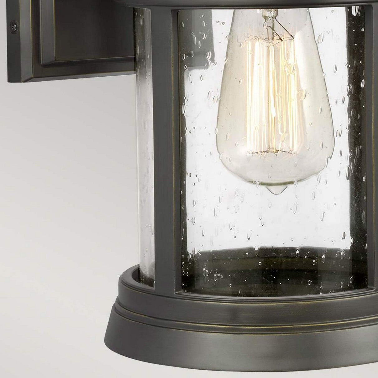 Close-up of the Newport 1 Light Outdoor Medium Wall Lantern in antique bronze, featuring a clear glass enclosure. Inside is a lit vertical Edison bulb showcasing its visible filament. The glass, adorned with water droplets from recent rain, adds to its coastal lighting charm.