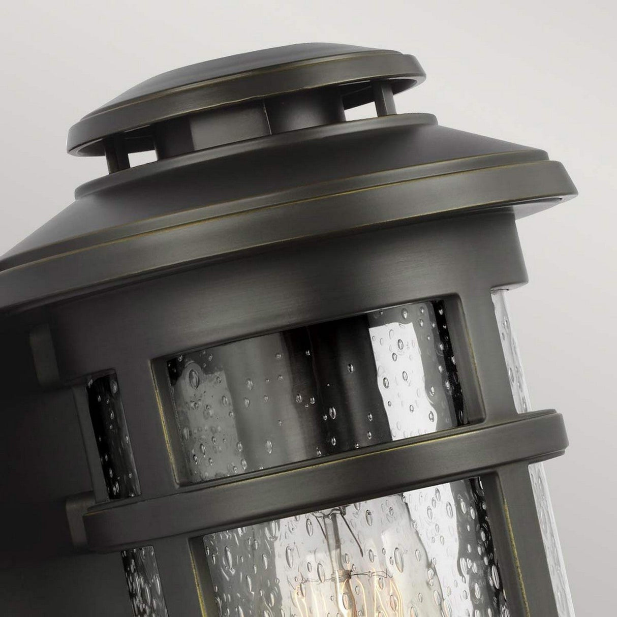 A close-up view of the Newport 1 Light Outdoor Small Wall Lantern in antique bronze, showcasing its clear glass panels adorned with visible raindrops. This coastal-style fixture encases a light bulb within, exuding a vintage and elegant charm.