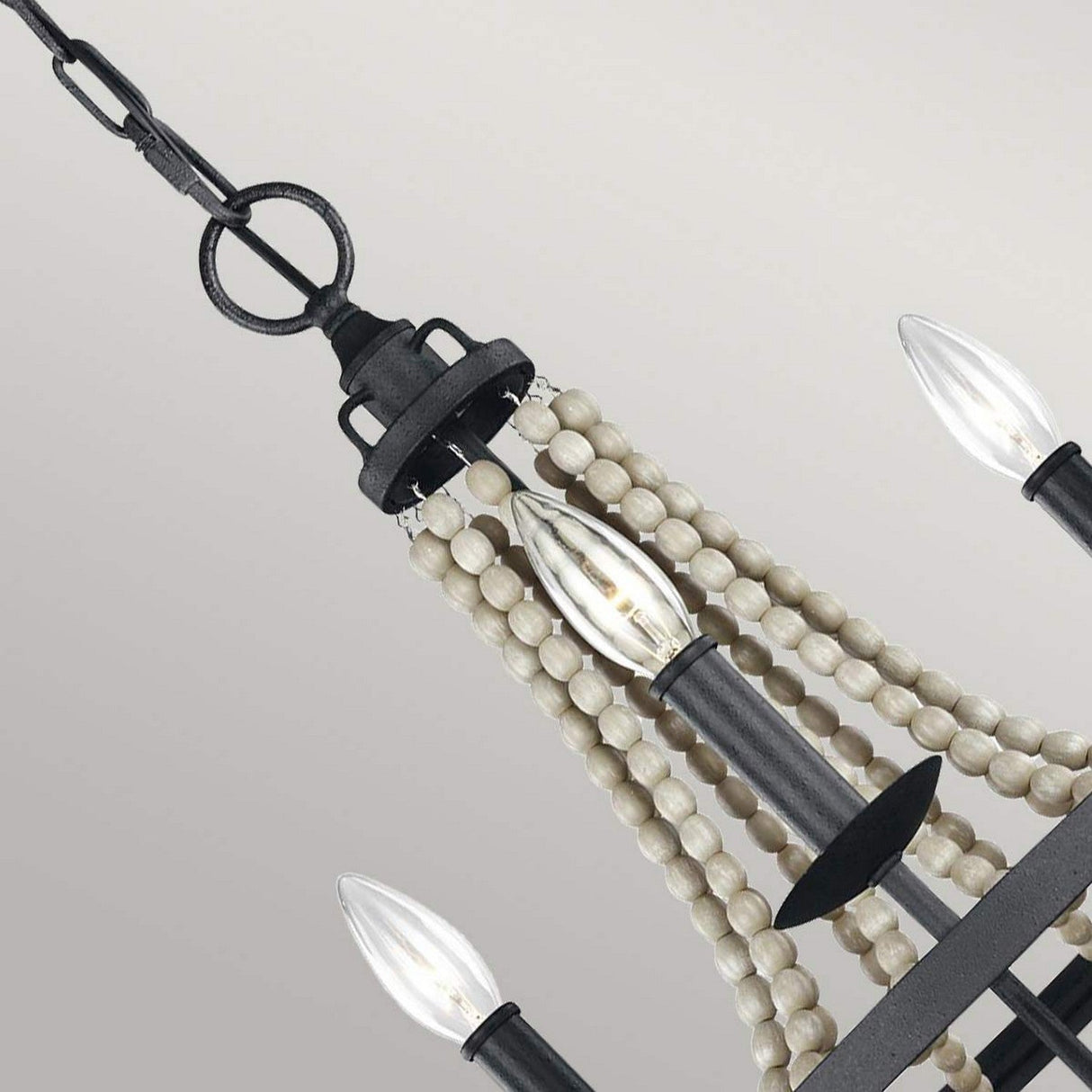A close-up view of the Nori 3 Light Chandelier - Dark Weathered Zinc With Driftwood Grey showcases its candle-shaped bulbs. The design includes strands of white beads elegantly draping from a dark weathered zinc frame, which is accented by driftwood grey tones. This piece artfully combines rustic and modern elements against a simple backdrop.