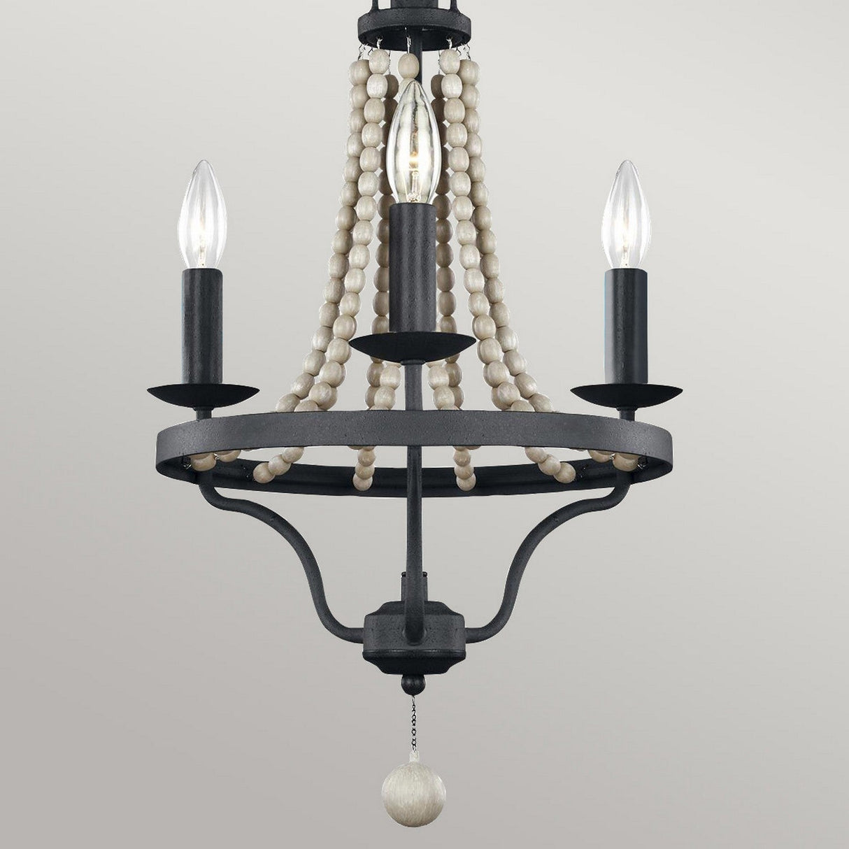 The Nori 3 Light Chandelier - Dark Weathered Zinc With Driftwood Grey showcases a sophisticated dark metal frame with candle-shaped bulbs and is beautifully accented with wooden bead strands. Its design is elegantly completed by a pull chain featuring a wooden knob, all highlighted in an exquisite Driftwood Grey finish.
