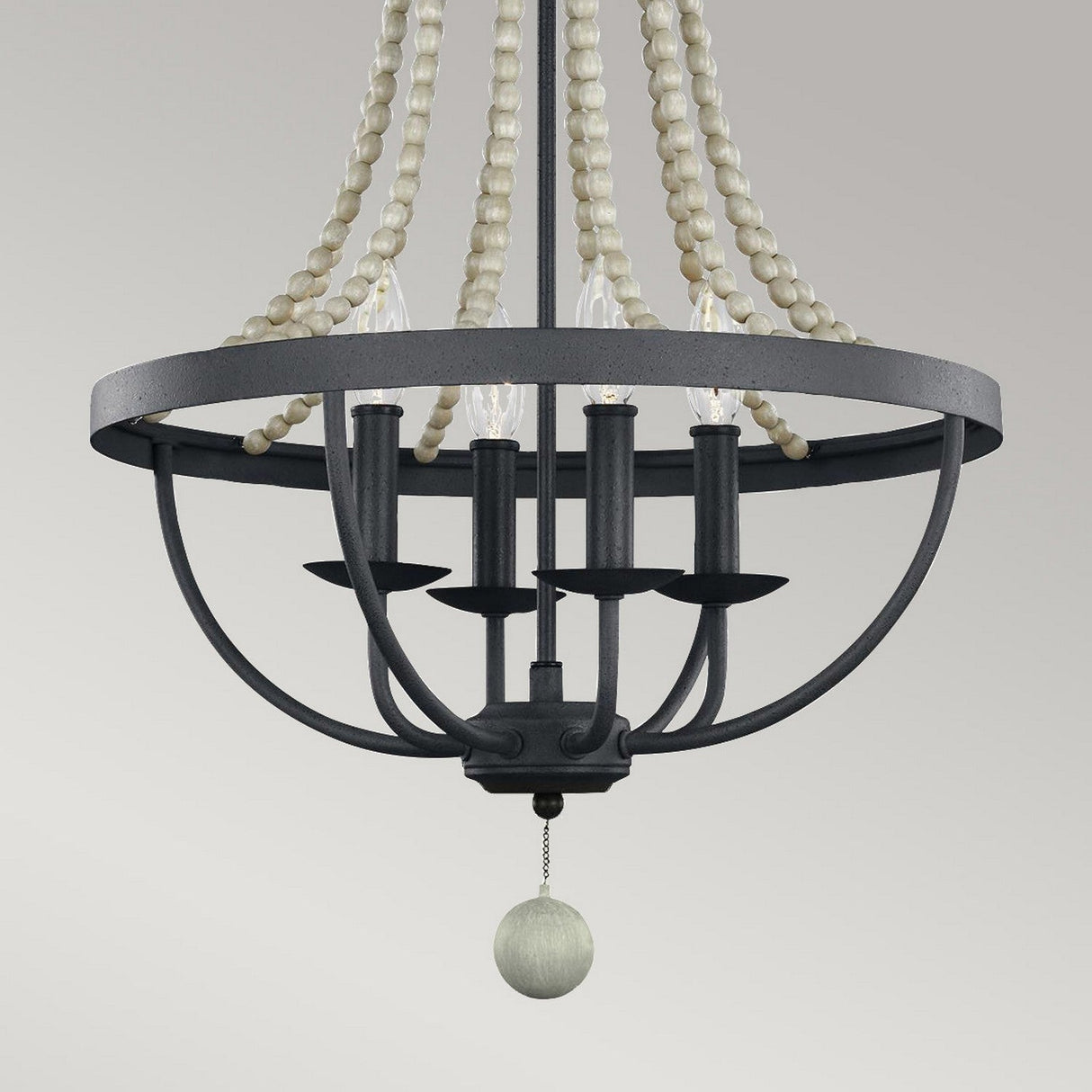 The Nori 4 Light Chandelier - Dark Weathered Zinc With Driftwood Grey features a striking black metal frame adorned with candle-style bulbs and graceful white beaded strands. A centrally positioned driftwood grey wooden sphere enhances the design, complementing its elegant appearance against a simple light gray backdrop, offering a touch of timeless charm.