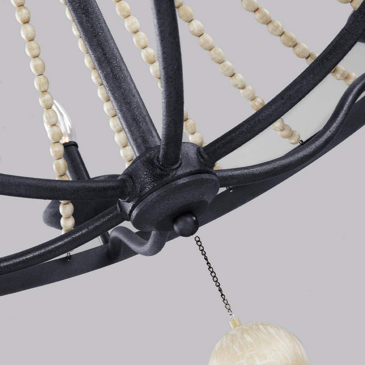 A close-up view of a Nori 6 Light Chandelier - Dark Weathered Zinc With Driftwood Grey reveals its dark weathered zinc arms intertwined with vertical strings of driftwood grey beads. A round bead is centrally suspended by a small chain, set against a plain gray background.