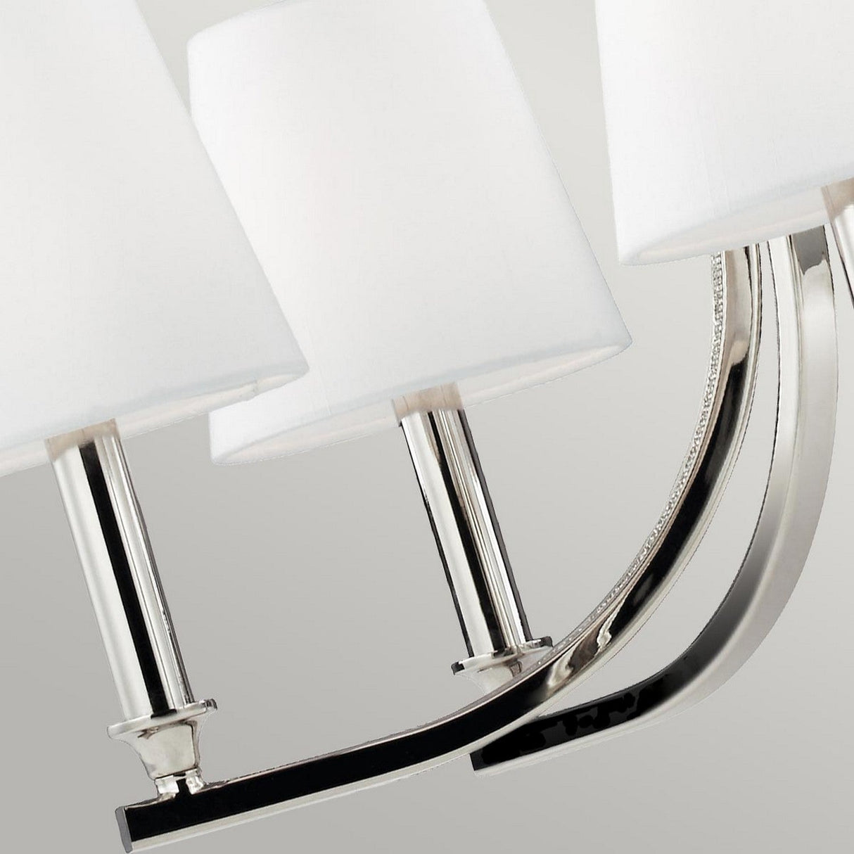 Close-up of the Pave 5 Light Chandelier showcasing its polished nickel finish with three elegantly curved chrome arms and white cylindrical lampshades. The sleek design and neutral tones contribute to its elegant, contemporary appearance reminiscent of a minimalist chandelier.