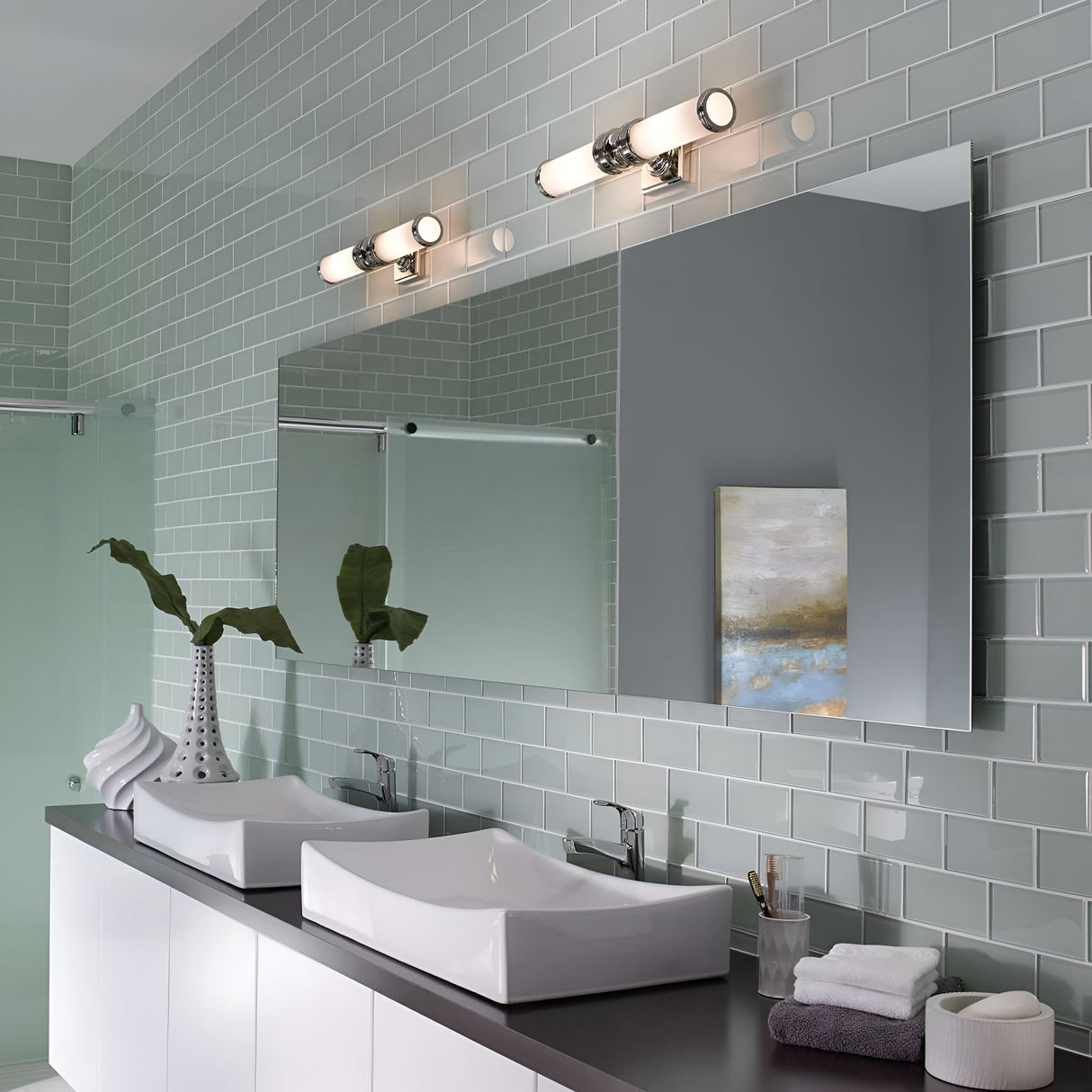 A modern bathroom showcases a large mirror above a double sink with white rectangular basins. The walls are adorned with light green tiles, perfectly complemented by the Payne-2 2 Light Wall Light in a polished chrome finish. A plant and decorative items enhance the countertop.