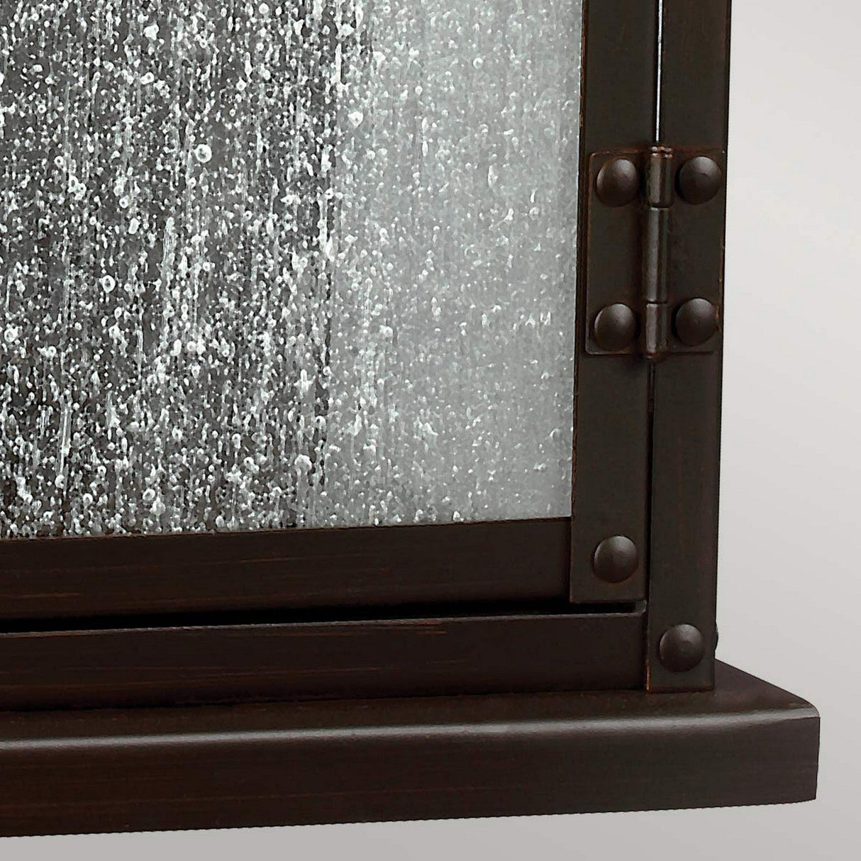 A close-up view of a textured, frosted glass panel in the **Pediment 2 Light Outdoor Medium Wall Lantern - Dark Copper** evokes the look of seeded glass panels. The speckled appearance resembles rain or condensation. The frame has a dark aged copper finish, showcasing visible hinges and corner details against a plain background.