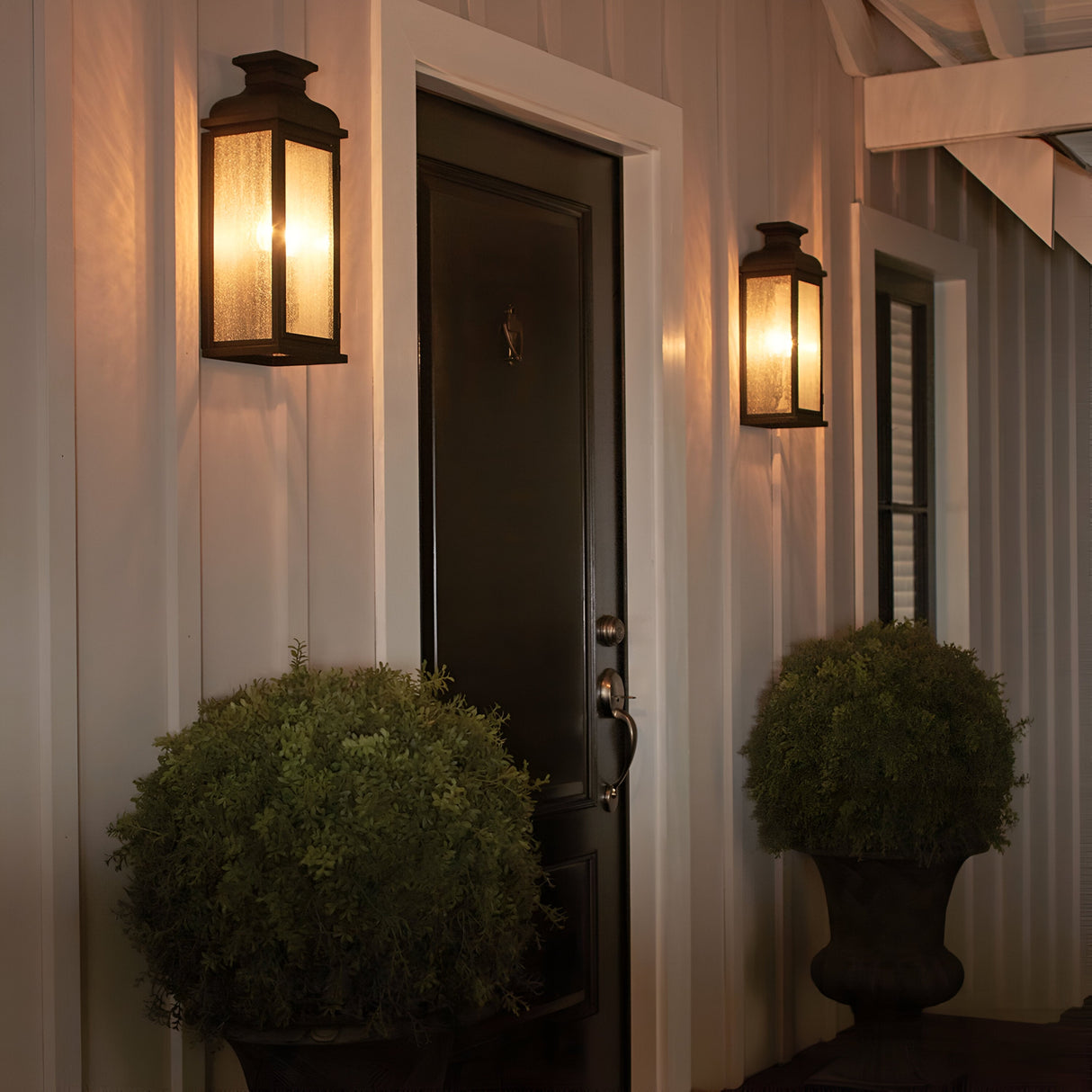 A cozy porch featuring a closed black door is framed by two Pediment 2 Light Outdoor Medium Wall Lanterns in dark copper. On either side of the door, two potted shrubs in dark planters rest against light-colored paneling, creating a warm and inviting ambiance complemented by the lanterns' seeded glass panels.