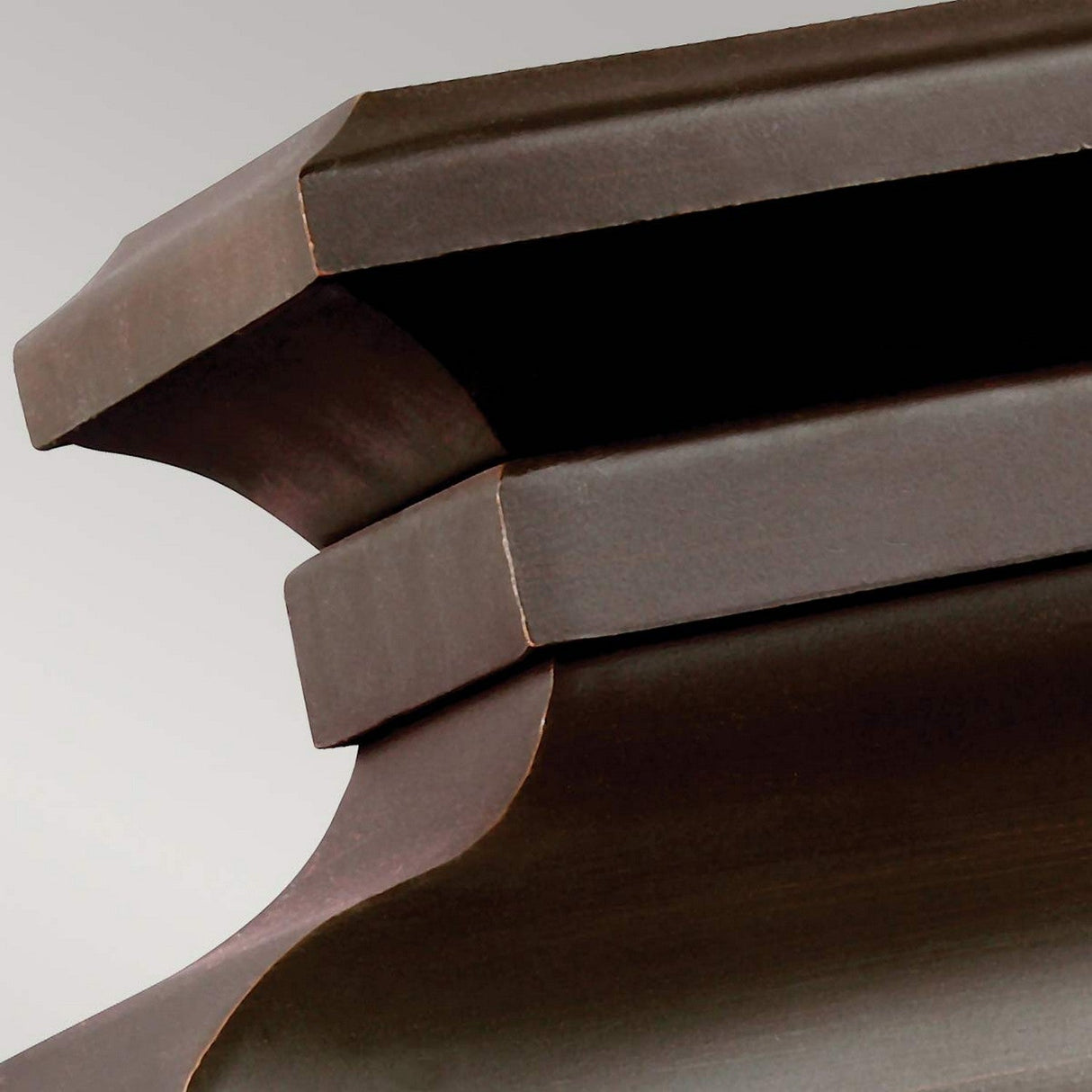 A close-up of the Pediment 2 Light Outdoor Small Wall Lantern in dark copper showcases its sculpted structure with curved, layered edges. The smooth finish accentuates its elegant craftsmanship and design, reminiscent of fine wooden furniture, highlighting the sophisticated seeded glass panels.