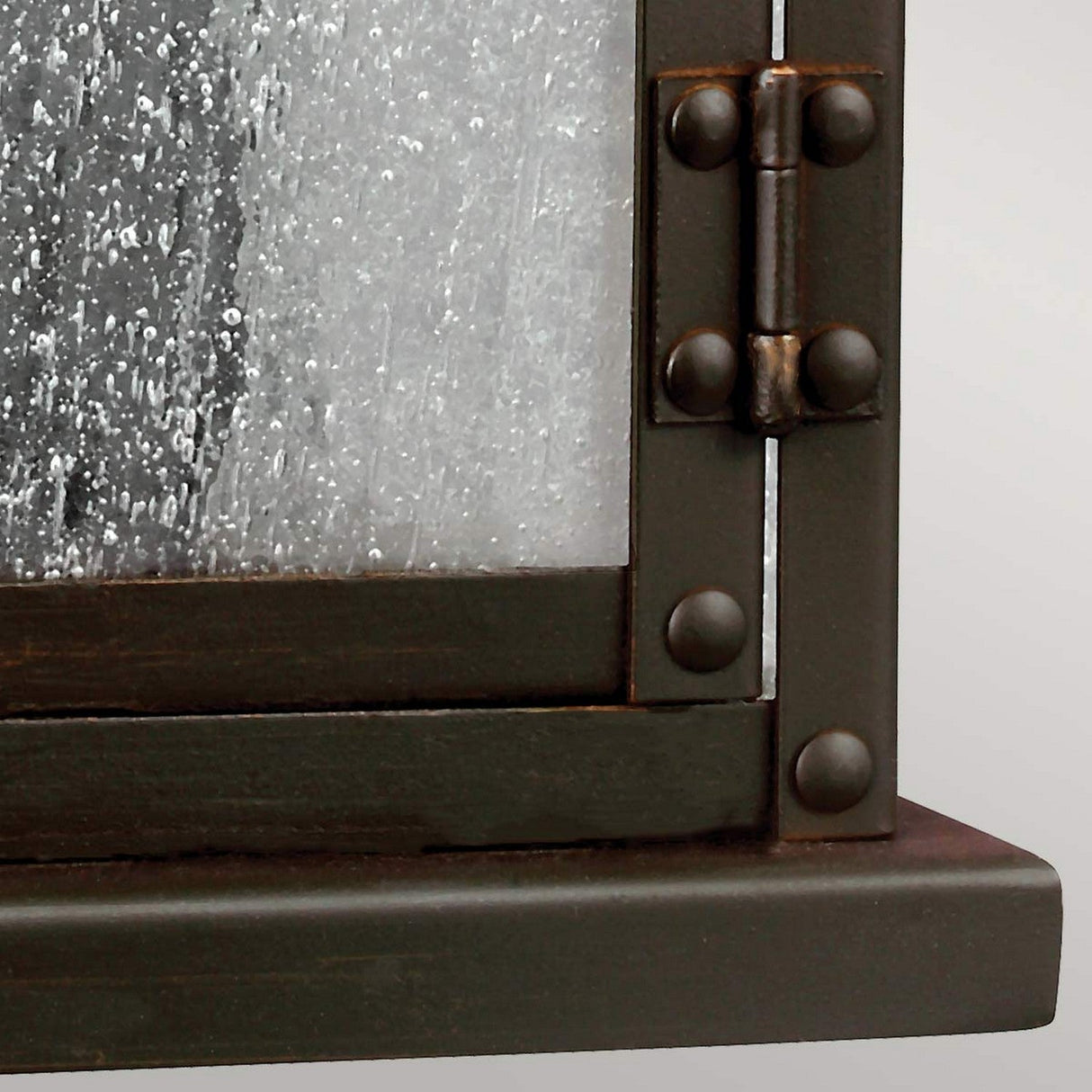 A close-up image showcases a frosted glass window with the appearance of seeded glass panels, reminiscent of water droplets for a blurred look. The frame features a dark aged copper finish, complete with visible rivets and a matte texture, similar to the design of the Pediment 2 Light Outdoor Small Wall Lantern in Dark Copper.