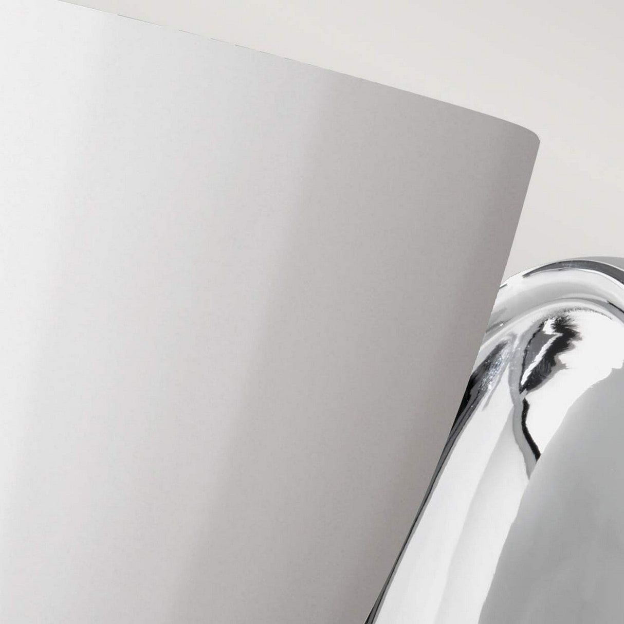 Close-up of a shiny, curved Perry 1 Light Wall Light in polished chrome next to a large, white, opal glass panel. The two surfaces create a striking contrast between the reflective metal and the matte white material, reminiscent of a sleek bathroom light fixture.