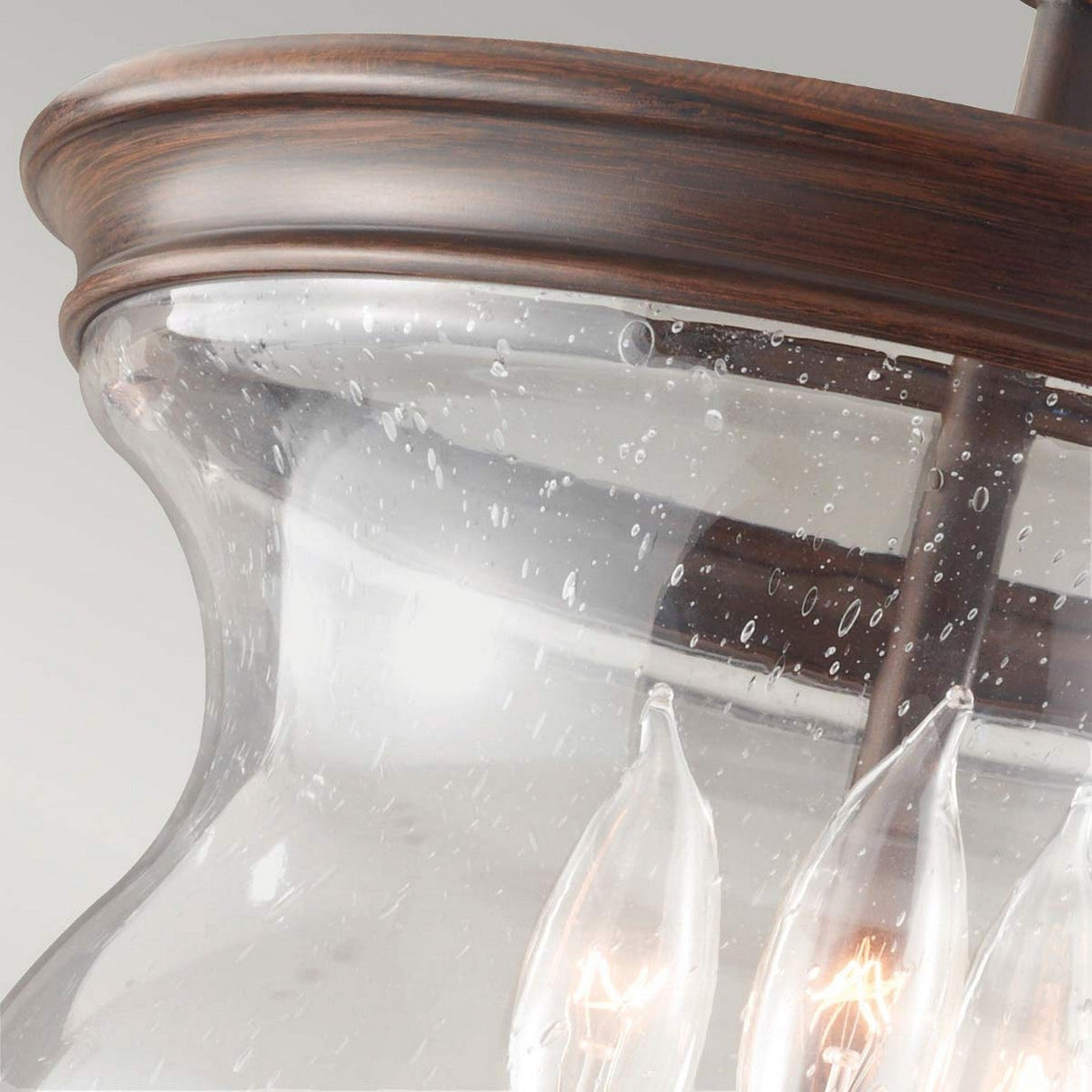 Product Description: A close-up view of the Pickering Lane 3 Light Semi Flush in Bronze showcases its glass pendant design, featuring a wooden rim and visible light bulbs within. The seeded glass shade provides a bubbled texture, emitting a soft glow that complements its rustic farmhouse style against a neutral background.