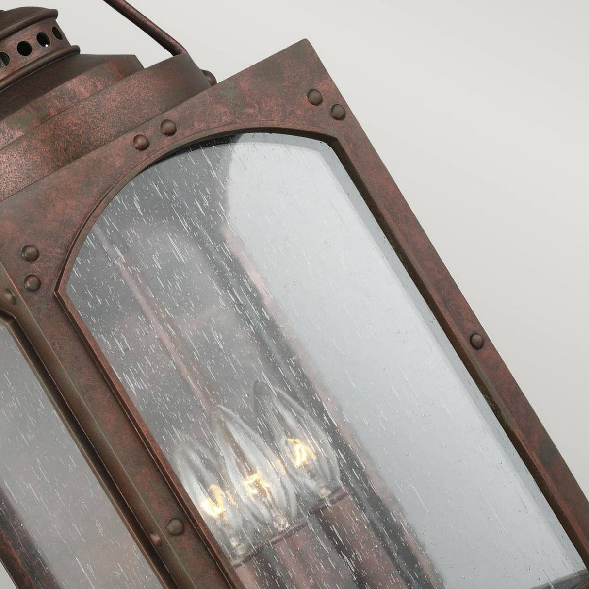 A detailed view of the Randhurst 3 Light Outdoor Large Wall Lantern - Copper reveals a rectangular glass pane with raindrop texture, housing multiple candle-style lights that emit a warm glow. The lantern features a copper oxide finish and riveted design, emphasizing its robust composite materials.