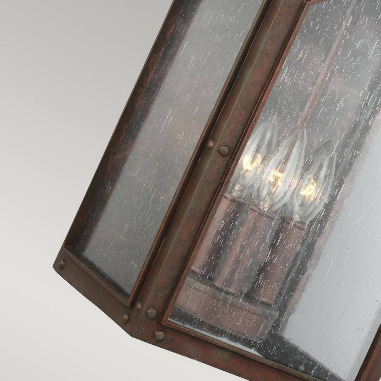 Close-up of the Randhurst 3 Light Outdoor Large Wall Lantern - Copper, showcasing its vintage-style design with a weathered metal frame in a copper oxide finish and clear, textured glass panels. Inside, three candle-like light bulbs emit a warm glow. The fixture hangs at an angle against a neutral background, highlighting its timeless elegance.