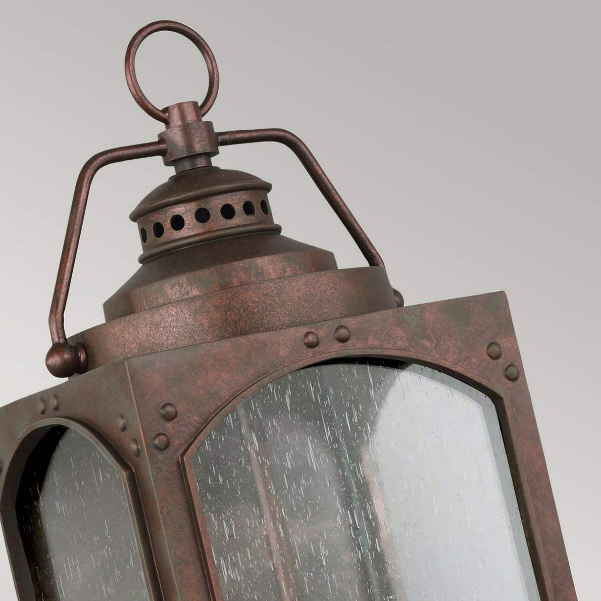 Close-up of the Randhurst 3 Light Outdoor Large Wall Lantern - Copper, featuring vintage bronze with clear glass panes, showcasing its weathered texture and intricate metalwork. The top includes a round handle, and it is enhanced with a copper oxide finish that radiates rustic charm against a neutral gray background.