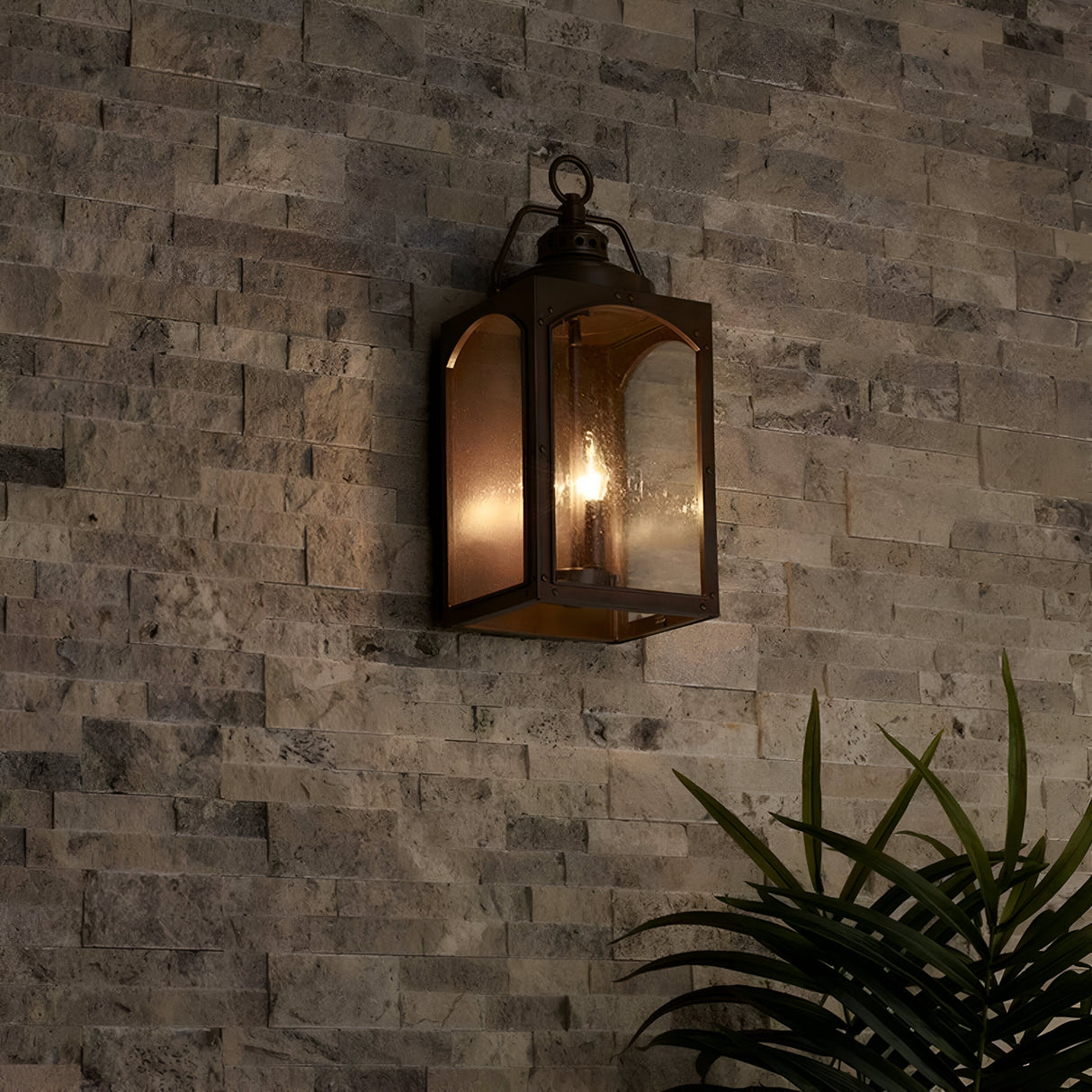 The Randhurst 3 Light Outdoor Large Wall Lantern - Copper is mounted on a textured stone wall, emitting a warm glow. A green plant partially appears in the lower right corner, adding a touch of nature to the scene.