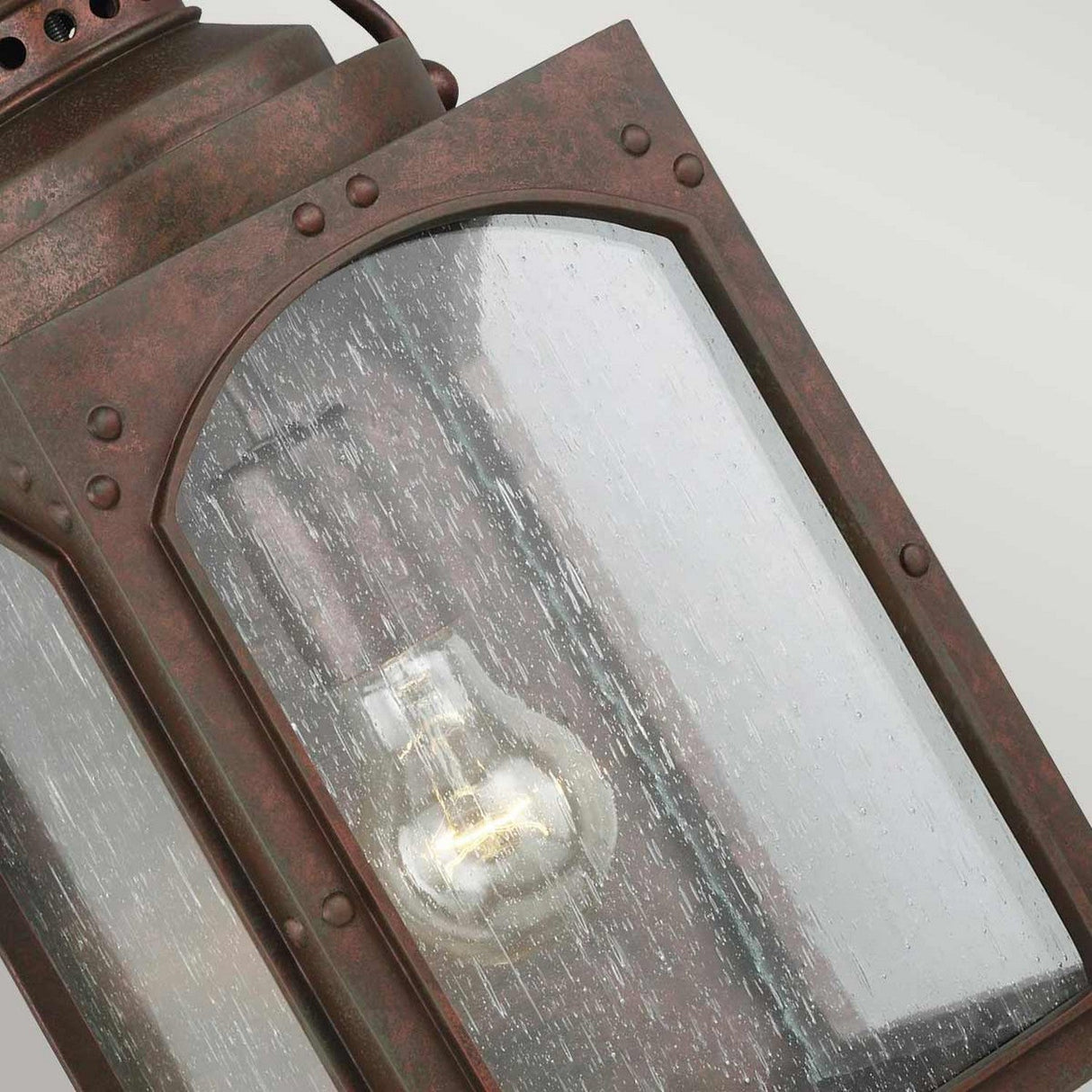 Close-up of the Randhurst 1 Light Outdoor Medium Wall Lantern - Copper, featuring a clear glass panel. The metal frame has a weathered bronze finish, reminiscent of copper oxide, with an exposed light bulb inside. The textured glass creates a water-like effect.