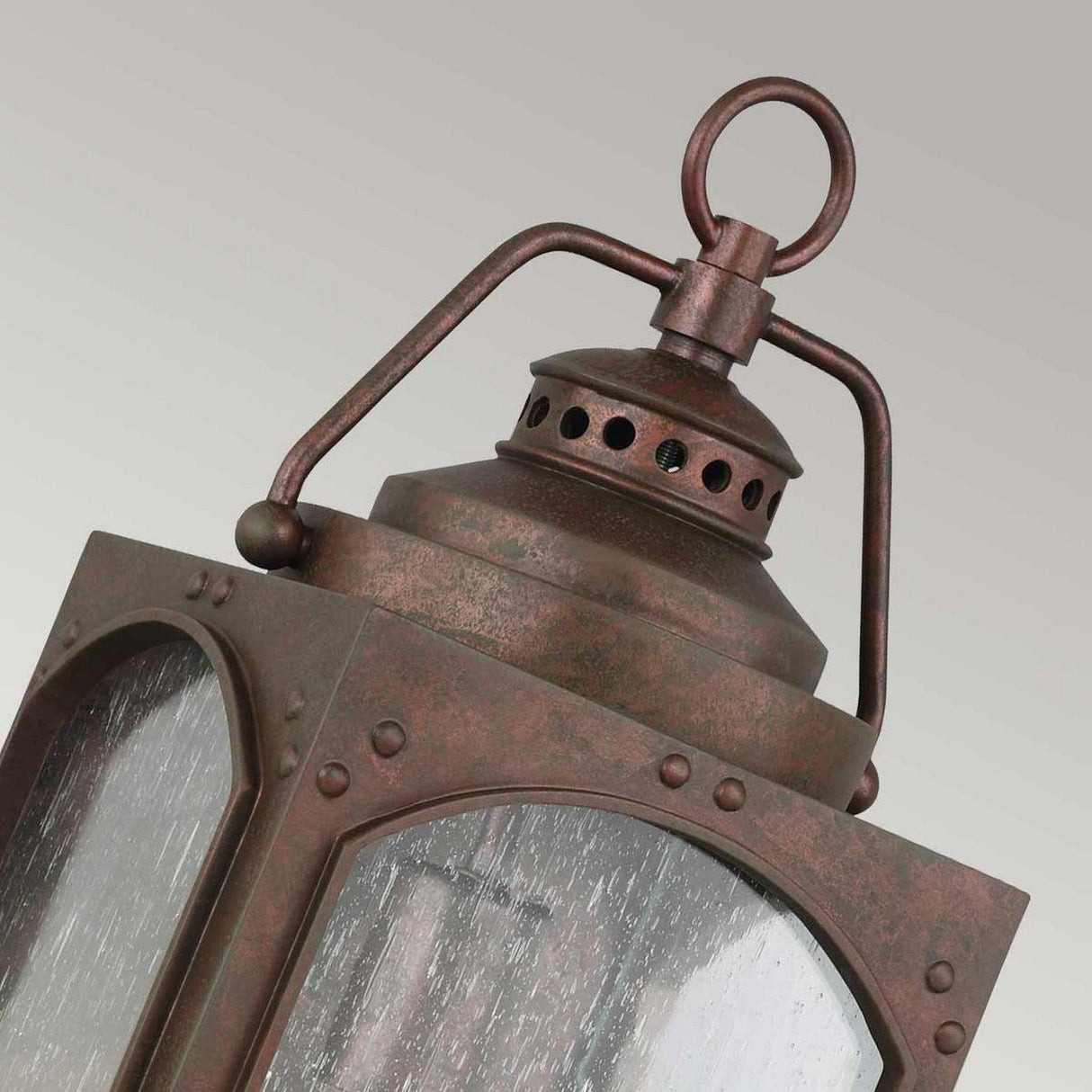 A close-up of the Randhurst 1 Light Outdoor Medium Wall Lantern, featuring a vintage style with a copper finish that exudes charm. It includes a ring handle on top, decorative perforations, and clear glass panels with visible texture, set against a plain gray background.