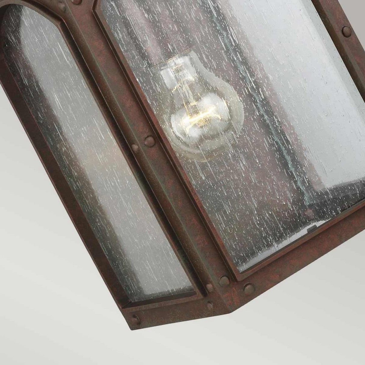 Close-up of the Randhurst 1 Light Outdoor Medium Wall Lantern – Copper, highlighting its vintage charm with a glowing light bulb inside. The lantern boasts a weathered metal frame with a copper oxide finish, and the clear, textured glass features a slightly speckled surface.