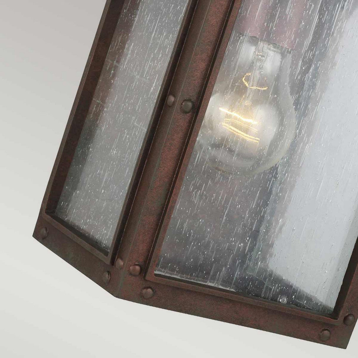 Close-up of a Randhurst 1 Light Outdoor Small Wall Lantern in copper, featuring a weathered metal frame and clear glass panels. The light bulb shines through the glass, showcasing a textured design that simulates rain droplets. This lantern offers a perfect blend of rustic charm and timeless elegance.