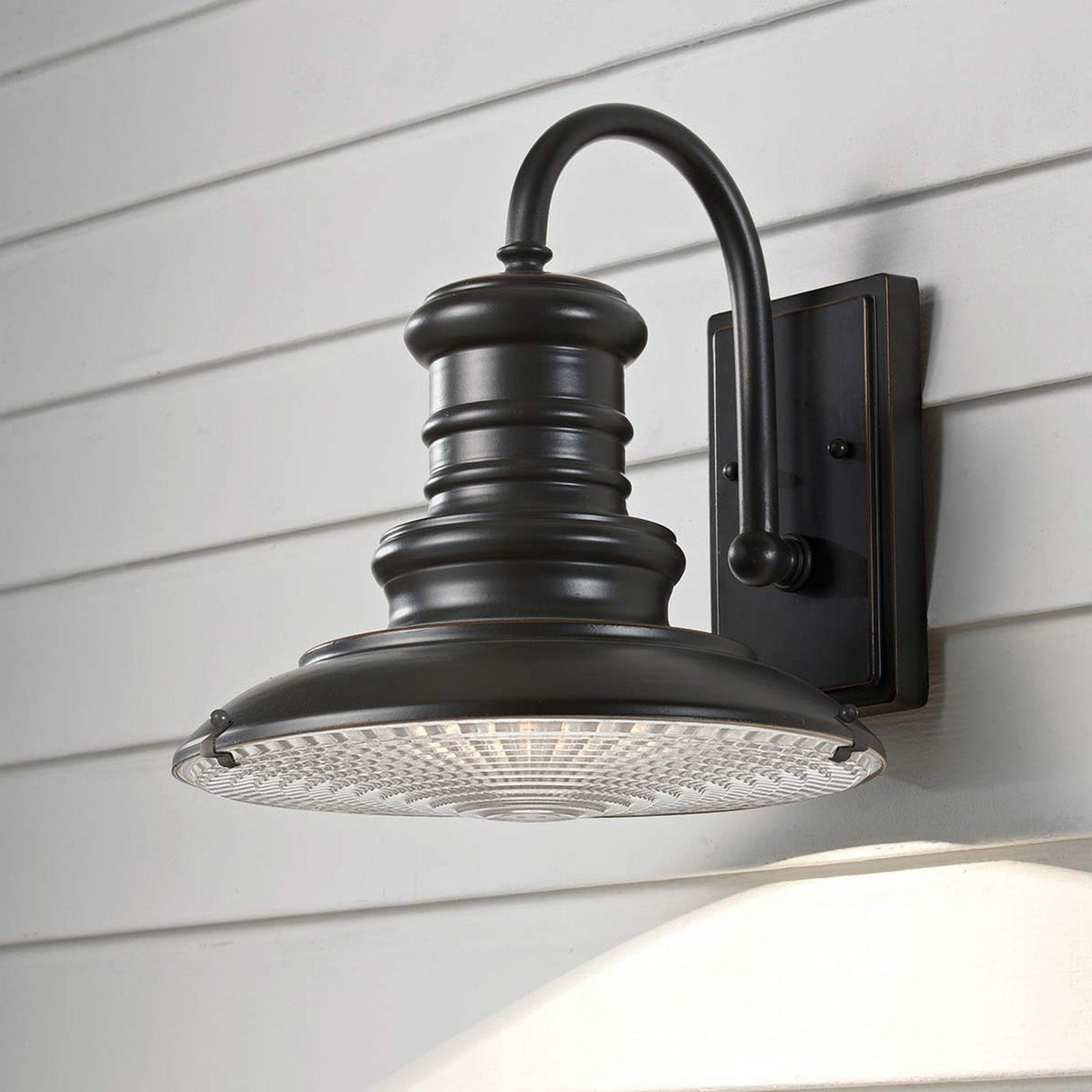 The Redding Station 1 Light Outdoor Medium Wall Lantern in dark bronze is elegantly mounted on a light gray, horizontally paneled wall. This weather-resistant fixture features a vintage style with a curved arm and wide, rounded shade topped with a textured glass cover.