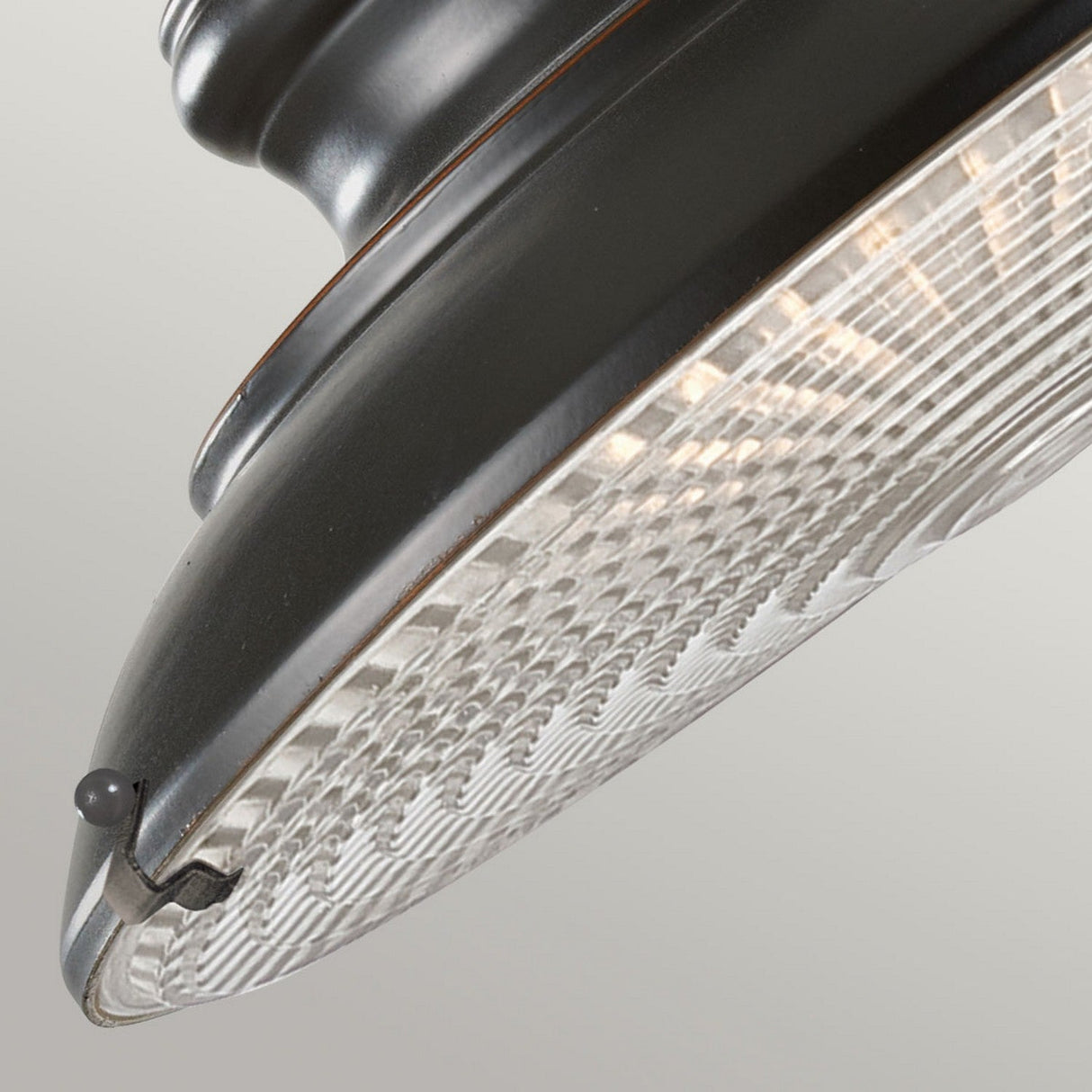 A close-up view of the Redding Station 1 Light Outdoor Medium Wall Lantern in dark bronze showcases its weather-resistant design with a textured glass cover that reflects light. Seen from below, the lantern features a small black knob at the edge and a subtle crack in the glass, set against a plain light gray background, highlighting its outdoor wall lantern style.