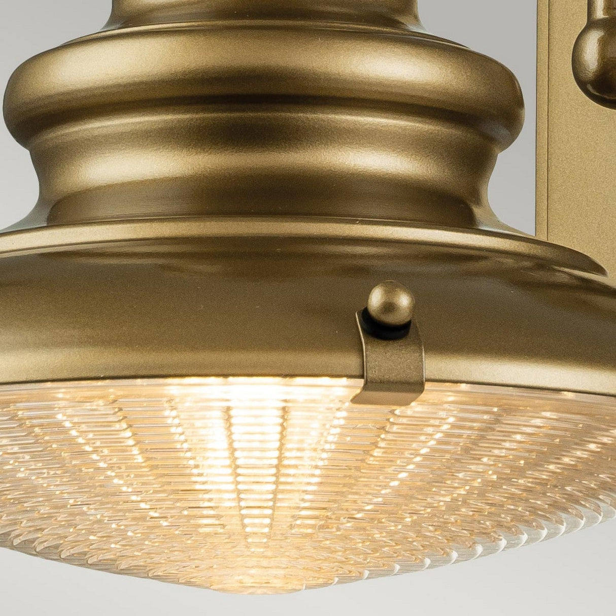 A close-up of the Redding Station 1 Light Outdoor Small Wall Lantern in bronze, featuring an industrial-style design with a prismatic glass shade. The illuminated light emits a warm glow through the textured glass. Its robust structure is suitable for wall or ceiling mounting.