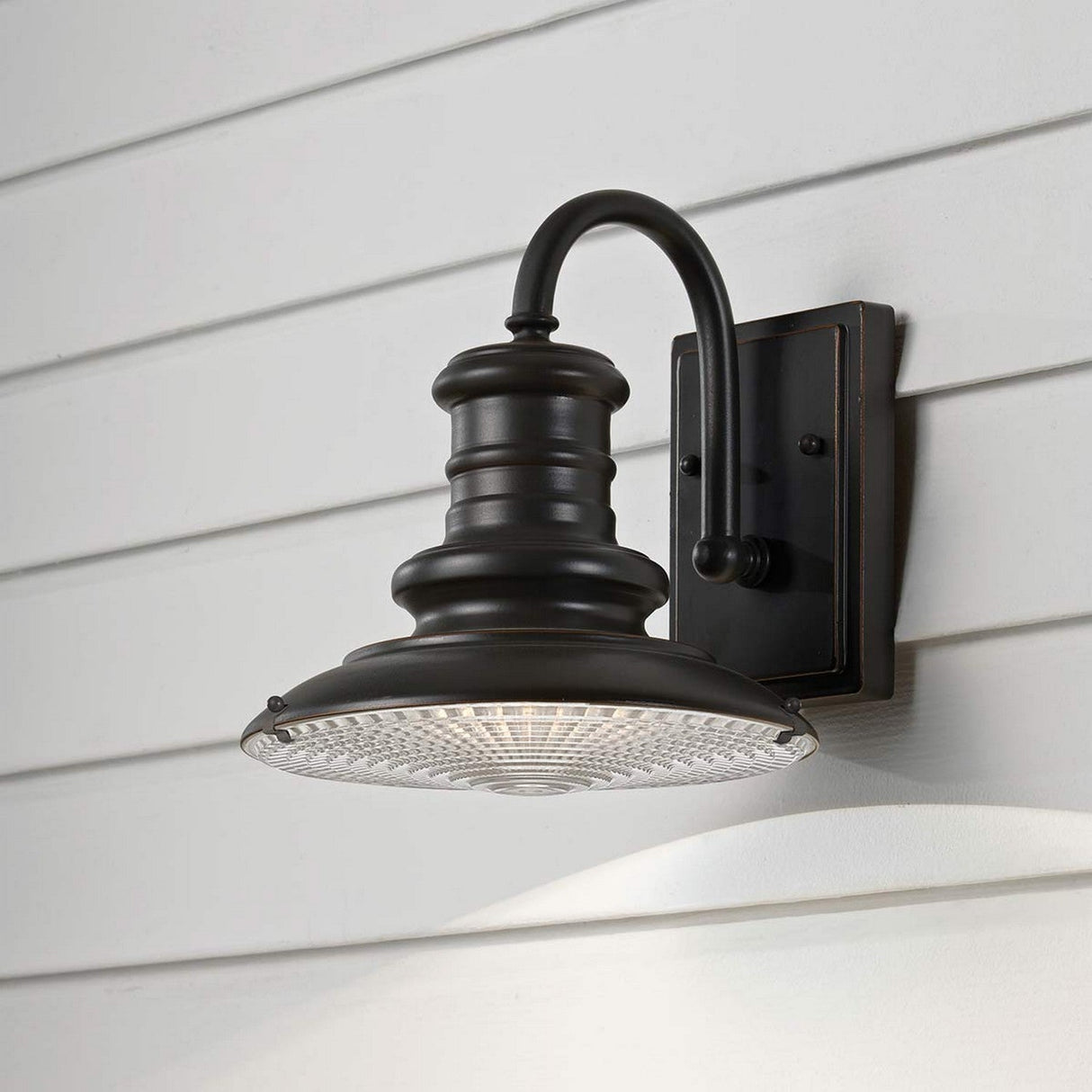 The Redding Station 1 Light Outdoor Small Wall Lantern, crafted in a dark bronze finish, is mounted on a white siding wall. It boasts an industrial-style design with a curved arm and a wide, round canopy. The prismatic glass shade adds texture and intrigue to the overall look.