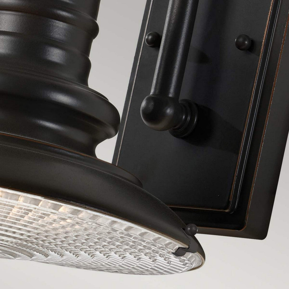 Close-up of the Redding Station 1 Light Outdoor Small Wall Lantern in dark bronze, showcasing a prismatic glass shade. This outdoor wall lantern has rounded edges and is mounted on a rectangular base with decorative knobs, providing a sleek, modern look.