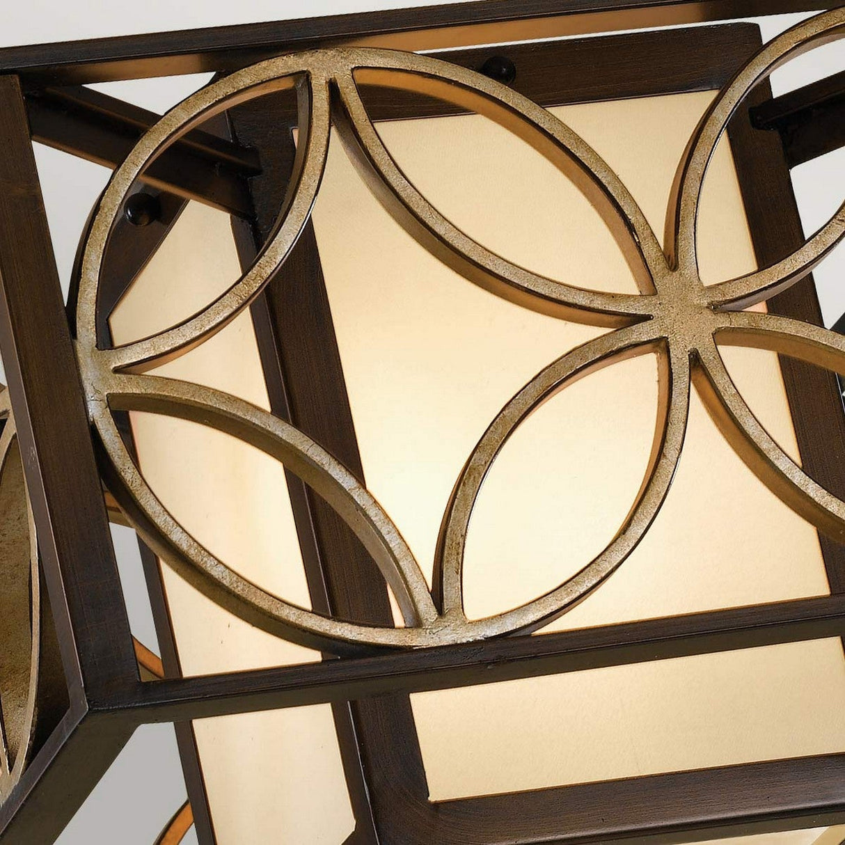 A close-up of the Remy 2 Light Flush in a Heritage Bronze finish with Parissiene Gold detailing highlights its geometric metal patterns layered over luminous frosted glass panels. The design casts a warm, ambient glow and presents an intricate circular motif that subtly echoes the elegance of French Arts and Crafts style.
