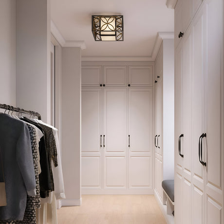A walk-in closet with white cabinets and wooden flooring showcases clothes hanging on a rail to the left and a cushioned bench built into the cabinetry on the right. The Remy 2 Light Flush, designed in French Arts and Crafts style, emits an elegant glow from above with its Heritage Bronze/Parissiene Gold finish.