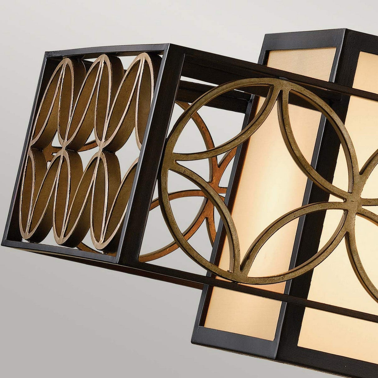 A close-up of the Remy 4 Light Pendant Chandelier in Heritage Bronze/Parisienne Gold showcases its interlocking circular designs. The illuminated panels, encased within rectangular frames, emit a warm glow.