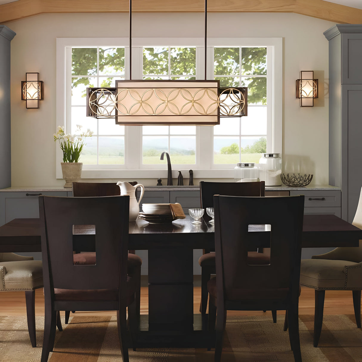 A modern dining room is highlighted by a dark wooden table surrounded by matching chairs, with the Remy 4 Light Pendant Chandelier in Heritage Bronze/Parissiene Gold serving as the central decorative fixture. A large window offers a scenic nature view, complemented by a vase of flowers, elegant glassware, and cabinetry in neutral tones.