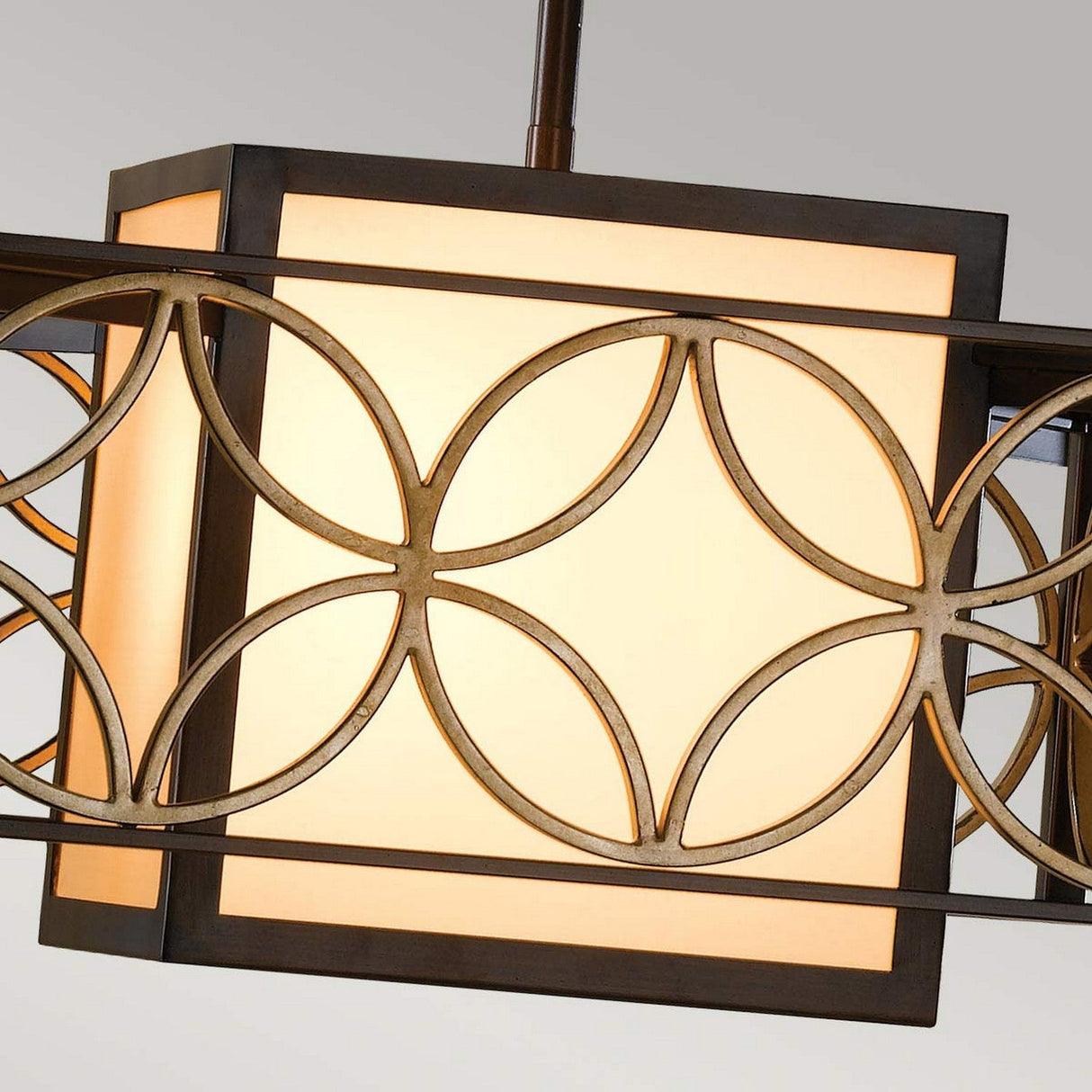 The Remy 1 Light Chandelier features a square pendant design with a warm glow, enclosed in a decorative bronze metal frame adorned with intersecting circular patterns that highlight artisan craftsmanship. Its Heritage Bronze finish, complemented by Parissiene Gold accents, adds an elegant touch against a neutral background.