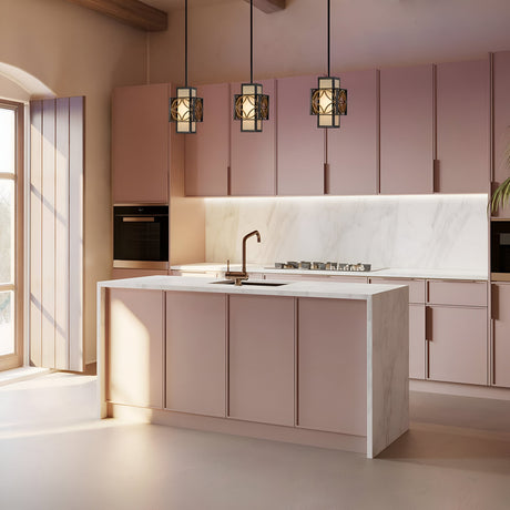 A modern kitchen showcasing pink cabinetry and a marble island boasts three Remy 1 Light Pendant Lights in Heritage Bronze/Parissiene Gold hanging above. Sunlight pours through a large window, brightly illuminating the spacious room where a built-in oven and stovetop can be seen.