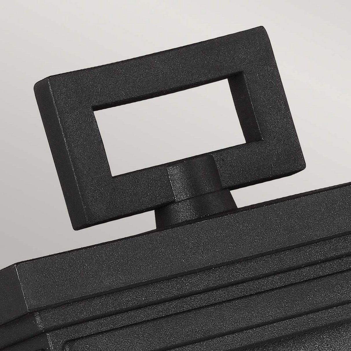 Close-up of the modern, geometric handle on the Roscoe 1 Light Outdoor Small Wall Lantern in Black. The weather-resistant handle boasts a hollow rectangular shape with a smooth finish. The background is a neutral, light gray color.