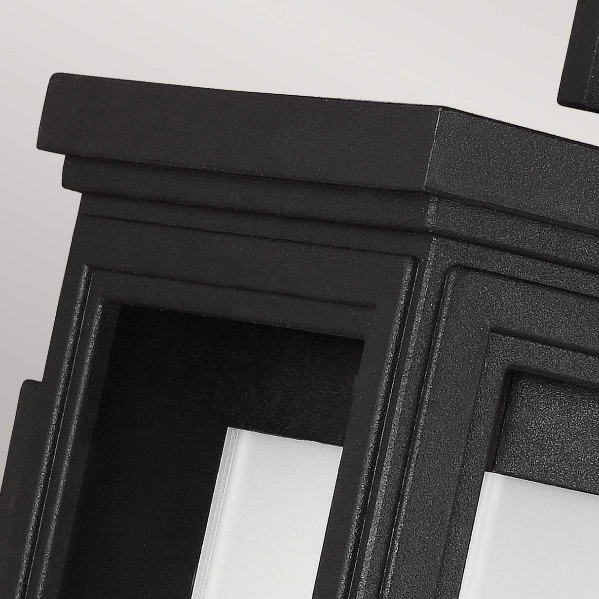 A close-up of the Roscoe 1 Light Outdoor Small Wall Lantern in black showcases its geometric, three-layered frame with sharp edges. Against a light gray background, the lantern exudes vintage charm while highlighting its structured and minimalist design. Its weather-resistant finish makes it perfect for an outdoor wall setting.