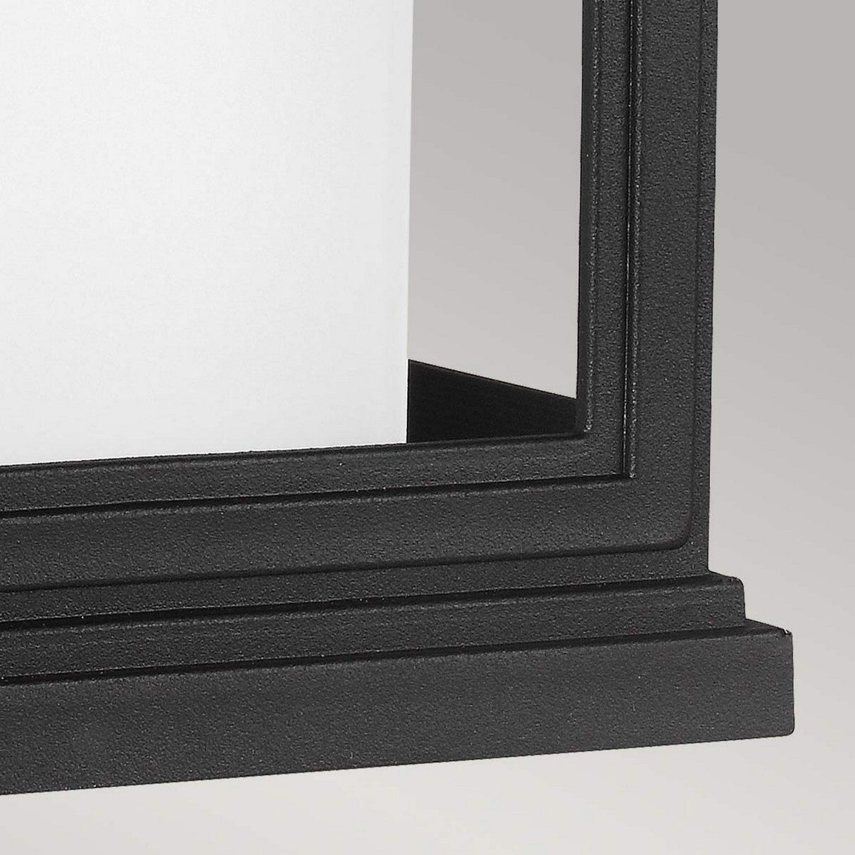A close-up of the corner of the Roscoe 1 Light Outdoor Small Wall Lantern - Black, featuring a black, rectangular frame with a white panel inside, exuding vintage charm against a light gray background.