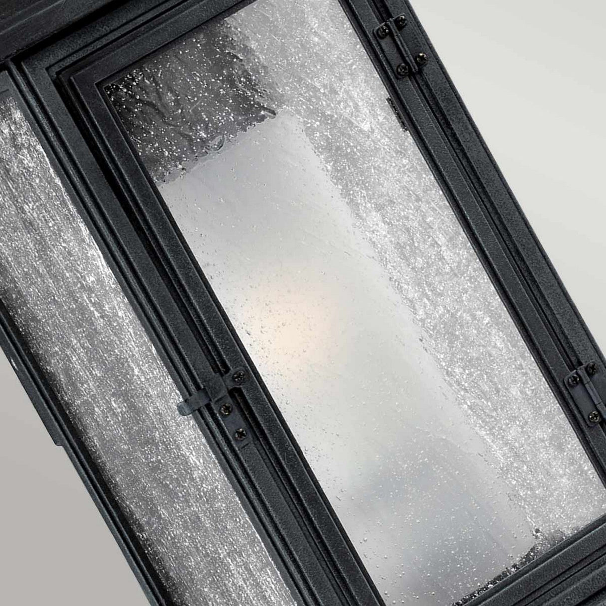 A close-up of a wet window with water droplets and frost surrounded by a dark frame reveals the blurred view outside, subtly illuminated by the gentle glow of the Shepherd 2 Light Outdoor Medium Wall Lantern in Dark Weathered Zinc. The classic Parisian design charm of the lantern enhances the dynamic perspective of the scene.