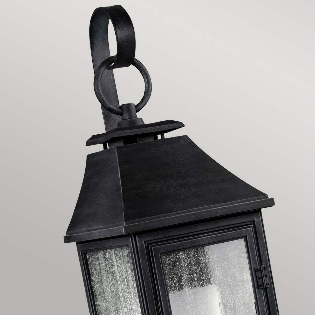 Close-up of the Shepherd 1 Light Outdoor Small Wall Lantern in Dark Weathered Zinc, ideal for vintage outdoor lighting. It features an angled design with a triangular top and loop handle. This weather-resistant lantern includes four glass panes and has a slightly weathered texture, set against a plain light gray background.