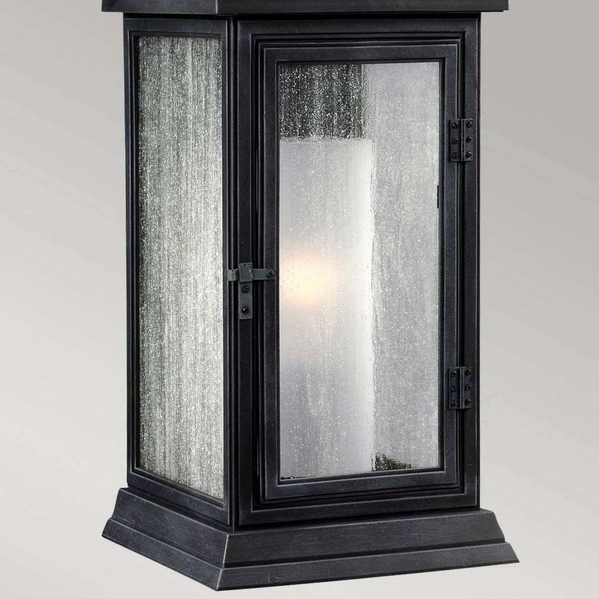 The Shepherd 1 Light Outdoor Small Wall Lantern in Dark Weathered Zinc showcases a vintage rectangular design with a single glass pane door and robust metal hinges. Its speckled textured glass softly illuminates the surroundings while being weather-resistant, all against a plain, light gray backdrop.