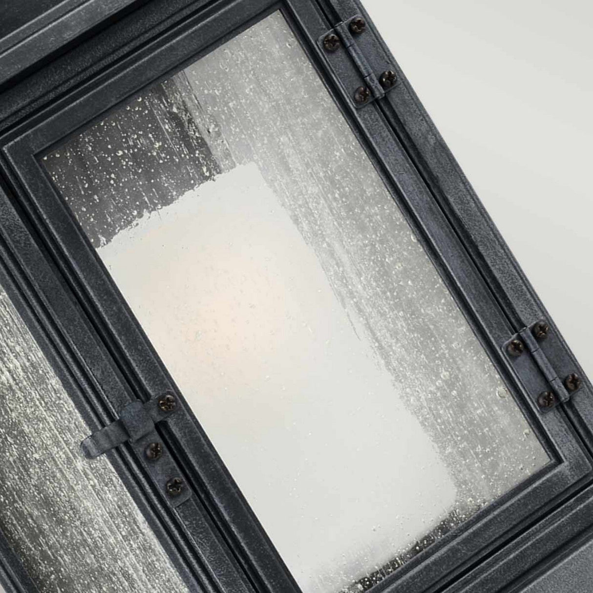 Close-up of the Shepherd 1 Light Outdoor Extra Large Wall Lantern in Dark Weathered Zinc, featuring a frosted glass design with a black metal frame, adorned with water droplets. A soft light glows through the glass, reminiscent of vintage street lighting, creating a warm and diffused illumination.