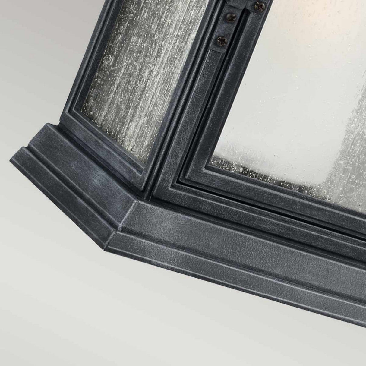 A close-up view of the Shepherd 1 Light Outdoor Extra Large Wall Lantern in Dark Weathered Zinc shows a black metal-framed glass panel with a textured surface. The corner design, featuring beveled edges, suggests its construction from weather-resistant composite material as part of a larger decorative architectural element.